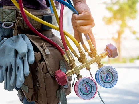 A person holds HVAC manifold gauges with attached hoses, wearing a tool belt and carrying work gloves, indicating air conditioning maintenance or repair work.