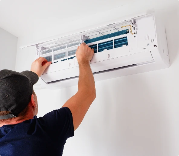 A person wearing a cap is inspecting or cleaning the filter of a wall-mounted air conditioning unit.