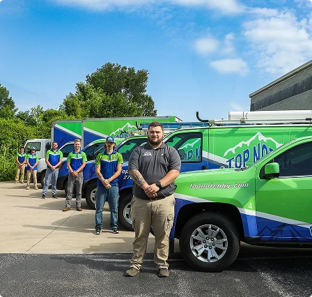 Five people stand in front of three branded service vehicles parked on a driveway, with trees and a building in the background.